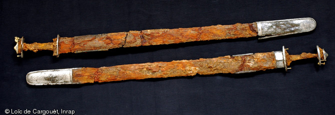 Épées provenant de deux tombes de  chefs francs , 525-550, Saint-Dizier (Haute-Marne), 2002.  Elles sont conservées dans leur fourreau en bois d'aulne terminé par une bouterolle en argent. L’épée (en haut sur la photo) comporte un pommeau en argent doré prolongé par deux anneaux. Le pommeau de l’autre épée possède une échancrure, attestant de la présence possible d’un double anneau, mais qui aurait été retiré. Signes distinctifs de ces tombes d'élites du VIe s., ces anneaux symboliseraient un lien d'homme à homme. 