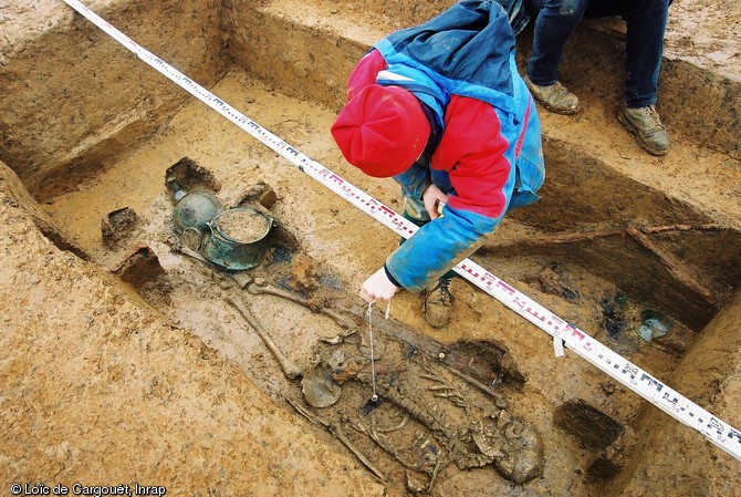 Relevé d'une des trois des riches tombes de Saint-Dizier (Haute-Marne), datées des années 525-550 et découvertes en 2002.  Le cercueil et le mobilier funéraire sont ici particulièrement bien visibles : aux pieds du défunt, on distingue les ferrures du cercueil en chêne, une bouteille en verre, un bassin et en chaudron en bronze.  En haut à droite du cliché, un angon et un fer de lance, déposés à l’origine sur le couvercle de la chambre funéraire et retrouvés affaissés à l’intérieur de l’excavation. 