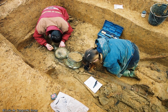 Fouille minutieuse d'une des riches tombes mises au jour à Saint-Dizier (Haute-Marne), 2002.  Datées des années 525-550, elles renferment un mobilier très riche, à l'image de ces deux récipients en bronze et de l'épée déposée le long du défunt. 