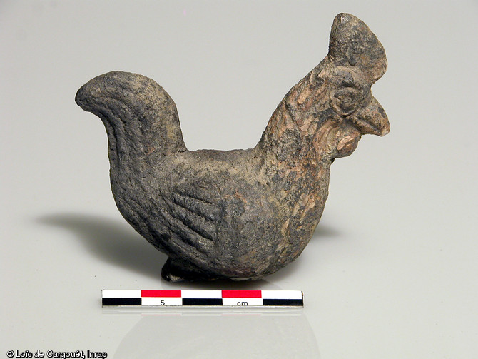 Figurine en terre cuite blanche représentant un coq, nécropole gallo-romaine de Pont-l'Evêque, Autun (Saône-et-Loire), 2004.  Cette petite représentation zoomorphe était destiné à être brûlée sur le bûcher funéraire, comme l'attestent les traces de combustion qu'on peut observer sur l'objet.