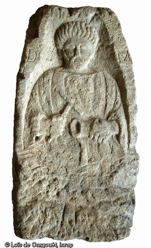 Stèle funéraire gallo-romain en arkose (hauteur 79 cm, largeur 25 cm), première moitié du IIe s. de notre ère, nécropole de Pont-l'Évêque, Autun (Saône-et-Loire), 2004.  Le personnage figuré est un artisan du métal, peut-être un dinandier : il tient un marteau et un gobelet dont le rebord est placé sur un support posé d'équerre. Son nom est illisible. Deux lettres seulement apparaissent, de part de d'autre du personnage : D et M, abréviations de Diis Manibus ( Aux dieux Mânes ). 