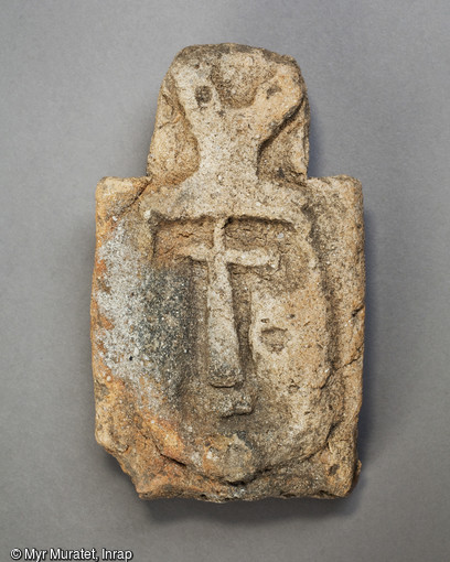 Tuile antéfixe en terre cuite , VIIe siècle, longueur 16,5 cm largeur 9 cm, découverte sur le site du Lac de la Médecinerie à Saran (Loiret) en 2011. L'identité du personnage représenté est probablement le Christ, caractérisé notamment les symboles apposés au-dessus de la tête sur le fronton. 