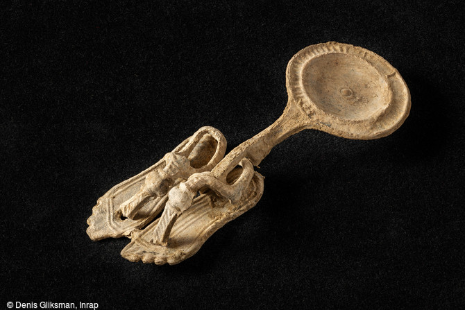 Miniature en plomb : paire de sandales savamment ornées accrochée à une patère (longueur : environ : 9 cm) découverte à Alba-La-Romaine (Ardèche), 2021. Un alignement spectaculaire de foyers de la Protohistoire ainsi qu'un ensemble funéraire antique ont été mis au jour. Ce dernier a livré un mobilier exceptionnellement bien conservé.
