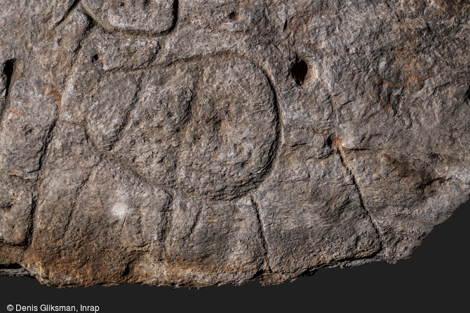 Vue de détail de la partie inférieure de la dalle de Saint-Bélec, découverte à Leuhan (Finistère). Forme patatoïde recoupée par des lignes verticales et ponctuée de petites cupules. Elle pourrait correspondre à une enceinte ou à un élément du relief.