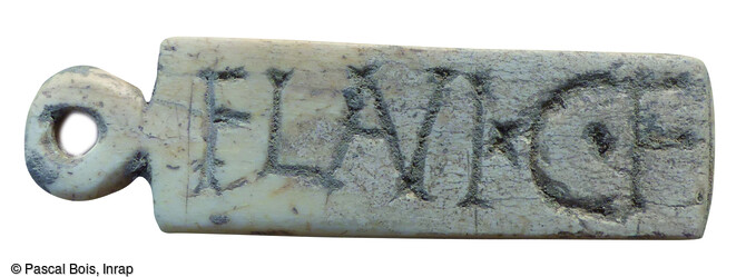 Détail d’un pendentif inscrit en os : FLAVI.C.F., signifiant Flavius fils de Caius. De riches vestiges d’un quartier urbain mis en place dès la Protohistoire (au IVᵉ siècle avant notre ère) et évoluant jusqu’à la fin du IIe siècle de notre ère ont été mis au jour de part et d'autres de la voie, à Nîmes (Gard), en 2021.