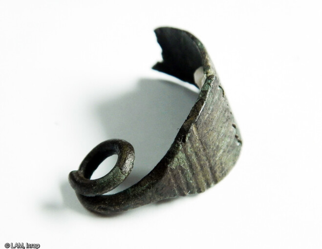 Fibule à ressort en alliage cuivreux découvert dans le paléochenal attribué à l'âge du Fer. Son arc est décoré au sommet de stries longitudinales et aux extrémités de stries transversales. L'avant de l'arc et l'ardillon ont disparus. Long. 2.6 cm ; larg. 1.2 cm ; poids. 2.1 gr
