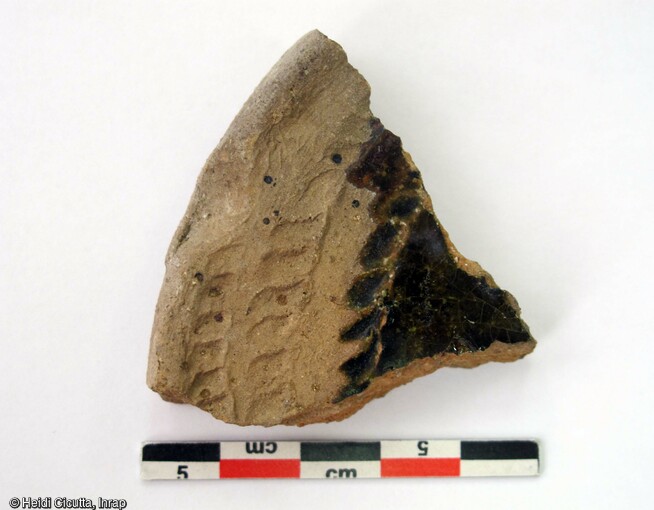 Fragment de couvercle en terre cuite glaçurée semi-grossière (glaçure verte sur cru) avec décor de rectangles à la molette, découvert dans une fosse du site et attribué au bas Moyen Âge (XVe s.).