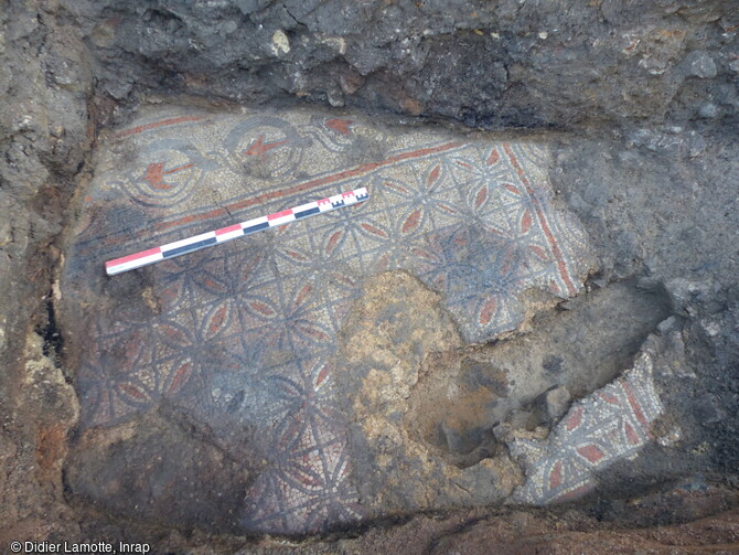 Vue de la mosaïque mise au jour dans le centre historique de Nevers (Nièvre) en 2023. Cette mosaïque se compose d'un panneau central constitué de rosaces en tesselles bleues et rouges sur fond blanc. Le bandeau qui borde le panneau central est conservé dans sa largeur, il montre un motif de tresse à deux brins associé à un symbole (hache ?) au centre des entrelacs. 