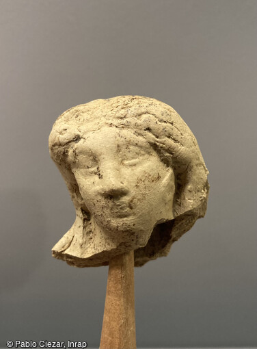 Fragment d'une figurine en argile blanche de l'Allier représentant une Vénus (1er-IIe s.) découvert lors de la fouille de l'École des Mines de Paris en 2023. Ce personnage féminin se distingue par une coiffure élaborée, caractéristique de l'époque, dite coiffure à la Vénus. Ce qui singularise cette coiffure, c'est son maintien en place grâce à un bandeau qui encercle la tête. Les mèches de cheveux, descendant le long des côtés, sont façonnées, formant un chignon de grand ampleur qui s'enroule vers l'extérieur. 