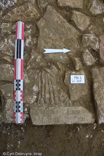 Fragment d'un monument funéraire mis au jour sur le parvis de la Basilique de Saint-Quentin (Aisne). Il s'agit d'un chanoine en prière décédé en 1496, portant l'aumusse sur le bras, en remploi dans les fondations du grenier de la Fabrique construit en 1717. 