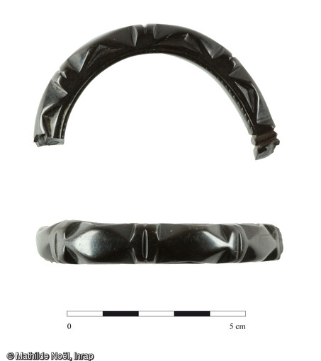 Brassard en jais sculpté datant de la fin de l'Antiquité (Bas Empire). L'origine de ce bijou est probablement à situer en Grande Bretagne dans le Yorkshire. Décor de triangle en creux sur les côtés donnant l'illusion d'une alternance de triangles. La vue sur le dessus est une alternance de losanges étirés séparés par deux petits bourrelets. Forme ovale incomplète de 7 cm de large, largeur bracelet de 0,8cm et épaisseur de 1,1 cm. Meung-sur-Loire (Loiret), Les Bouillants et la Maison Neuve
