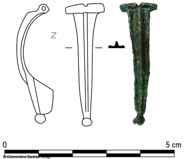 Dessin et photographie de la fibule en alliage cuivreux mise au jour dans le niveau de la voirie sur le site de Brumath (Bas-Rhin). Fortement corrodée, elle a fait l’objet d’un nettoyage mécanique et chimique et d’un recollage en laboratoire. Il s’agit d’une fibule dite « d’Aucissa », caractérisée par une charnière formée par le repli de l’arc vers l’extérieur, un porte-ardillon triangulaire plein, un arc rehaussé d’une arête médiane composée d’une ligne perlée et d' un bouton caudal. L’ardillon est néanmoins manquant. Ce type de fibule est particulièrement fréquent au début d