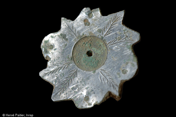 Fibule étamée avec décor de palmettes (diamètre 30 mm), Ier s. de notre ère, Neuville-sur-Sarthe (Sarthe), 2010.  Cet objet (une des nombreuses offrandes retrouvées sur le site) a été retrouvé dans un état de conservation surprenant : elle se présente ici telle qu’elle est sortie de terre.