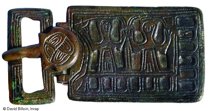 Plaque-boucle en bronze, fin VIe - début VIIe s., nécropole de Largillay-Marsonnay (Jura), 2001-2004.  Le décor met en scène deux personnages similaires, côte à côte, en position d'orants. Deux croix, situées aux angles du côté de la boucle, renforcent le caractère religieux de l'objet.     Photo publiée dans le numéro 20 de la revue de l'Inrap <a class= rte-link-ext  href= http://www.inrap.fr/archeologie-pr
