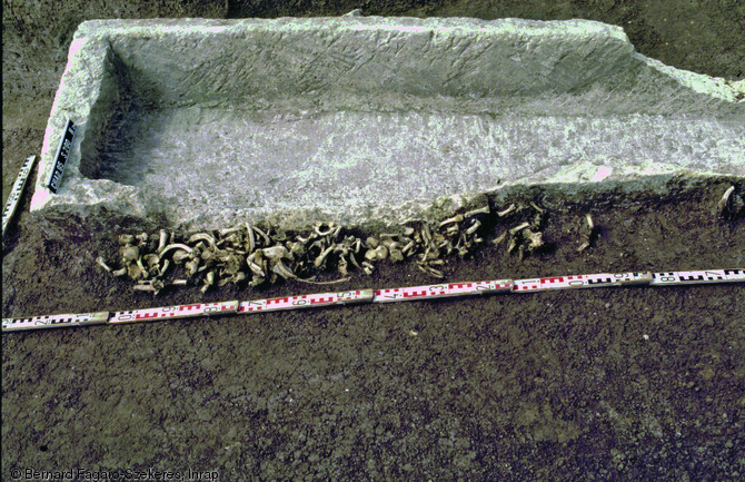 Sarcophage ayant abrité les os de plusieurs défunts, nécropole du haut Moyen Âge de Chadenac (Charente-Maritime), 1994.  Le long de la cuve est creusée une fosse contenant des ossements sans connexion, comprenant une large majorité de petits os : le sarcophage a été vidangé à plusieurs reprises afin d'accueillir de nouvelles sépultures, seuls les os de grande taille ont été laissés à l'intérieur.  Photo publiée dans le numéro 29 de la revue de l'Inrap <a class= rte-link-ext  href= http://www.inrap.fr/archeologie-preventive/Recherche-scientifique/Archeopages/Les-numeros/28-29-30-