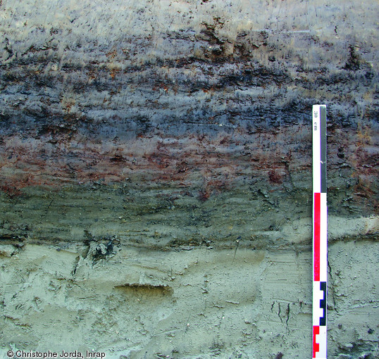 Détail d'une coupe stratigraphique réalisée dans des niveaux tourbeux sur le site de Marsillargues (Hérault), 2009.  C'est dans la couche brun-foncé à noire (au centre) qu'ont été trouvées les traces d'une occupation humaine s'étalant du IXe au XIe s., moment où l'environnement passe d'un milieu laguno-marin à un milieu terrestre.     Photo publiée dans le numéro 30 de la revue de l'Inrap <a class= rte-link-ext  href= http://www.inrap.fr/archeologie-preventive/Recherche-scientifique/Archeopages/Les-numeros/28-29-30-HS02-2010/Numero-30/Dossier-Homme-et-litto