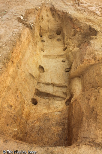 Cave du premier âge du Fer avec rampe d'accès visible à l'arrière-plan, vers 500 avant notre ère, Kervouyec (Finistère), 2011.  Les structures circulaires en creux sont les creusements laissés par des poteaux de bois qui soutenaient le plancher. 