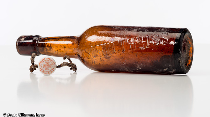 Bouteille de bière en verre brun gravée au nom de la brasserie  Schultheiss  de Berlin, avec son bouchon en porcelaine marqué  Schultheiss-Brauerei  autour d'une croix de Malte. Découverte en surface d'une fosse allemande datant de la Grande Guerre à Saint-Etienne-sur-Suippe (Marne), 2014.