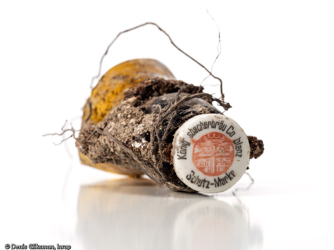 Col de bouteille de bière avec son bouchon en porcelaine au nom de la brasserie  König sbacherbräu Coblentz Schutz-Marke (Brasserie du ruisseau du roi, Coblence), avec en son centre probablement un blason aux armes de Coblence. Découvert en surface d'une fosse allemande datant de la Grande Guerre à Saint-Etienne-sur-Suippe (Marne), 2014. 