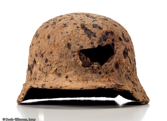 Casque en acier allemand (Stahlhem), modèle 1935, d. 25 cm et h. 25 cm, perforé de trois impacts probablement dus à des éclats d'obus, dont un frontal plus important. Datant de la Bataille de Normandie et découvert dans une fosse à Soumont-Saint-Quentin (Calvados), 2008. 