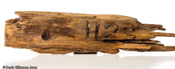 Fragment du dessus d'un couvercle de munitions en bois (L. 25 cm), d'origine allemande, sur lequel est indiqué, avec un marquage à chaud, le calibre et le type de pièce d'artillerie employée ;  H  pour  Haubitze  (obusier)  2(3 ou 9?). La ville de Tilly-sur-Seulles, fut le théâtre de violents combats, pendant l'opération Perch menée du 7 au 15 juin 1944 par les troupes du XXXe Corps britannique contre la  Panzer Lehr Division  allemande. Découvert dans une fosse à Tilly-sur-Seulles (Calvados), 2014.