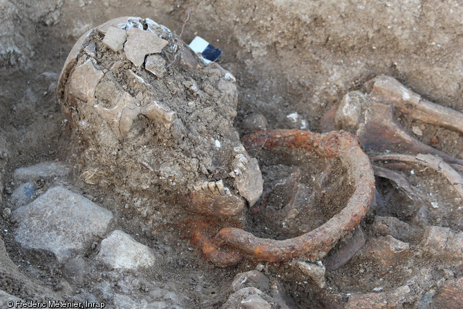 Vue du détail de la sépulture 2073 en cours de fouille. Individu masculin inhumé en décubitus, entravé au cou et à la cheville droite, découvert dans une nécropole antique à Saintes (Charente-Maritime).  L'entrave du cou se compose de deux parties semi-circulaires et de section circulaire, terminées par un  oeil  à l'une des extrémités, ce qui permet la mobilité des deux brins. 